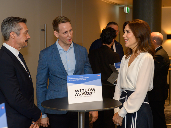 Folk i forretningstøj står og snakker ved et højt bord med et »WindowMaster«-skilt ved en konference eller et netværksarrangement.