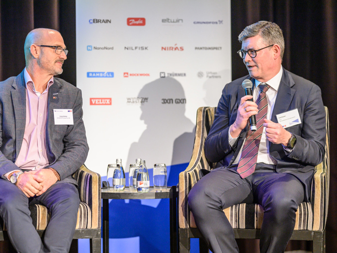 To mænd i jakkesæt, der diskuterer på scenen, med en mikrofon, vandflasker og et sponsorplakat i baggrunden.