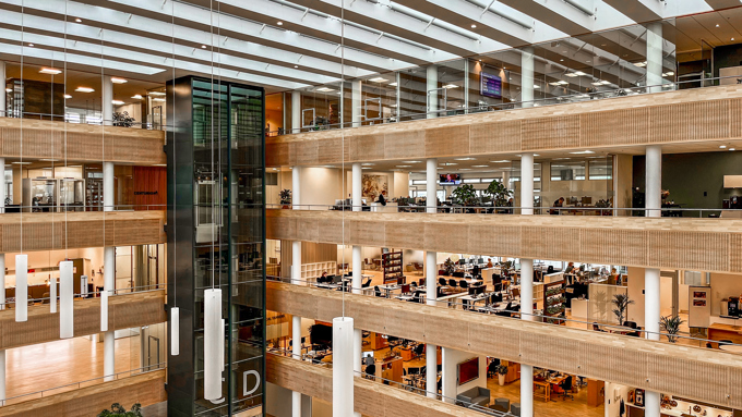Moderne kontorindretning med flere etager, elevator og store åbne rum fyldt med grønne planter og arbejdsstationer.