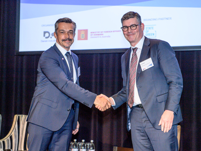 To mænd i jakkesæt, der giver hinanden hånden ved en formel begivenhed med en præsentationsskærm og vandflasker i baggrunden.