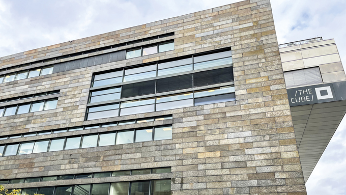 Moderne bygningsfacade med vandrette stenpaneler og store vinduer, med »THE CUBE«-skiltning på siden.