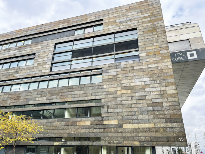 Moderne bygningsfacade med vandrette stenpaneler og store vinduer, med »THE CUBE«-skiltning på siden.
