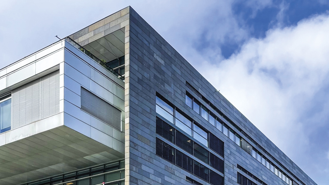 Moderne bygning med en elegant stenfacade og store glasvinduer under en delvis overskyet himmel. Træer og biler er i forgrunden.