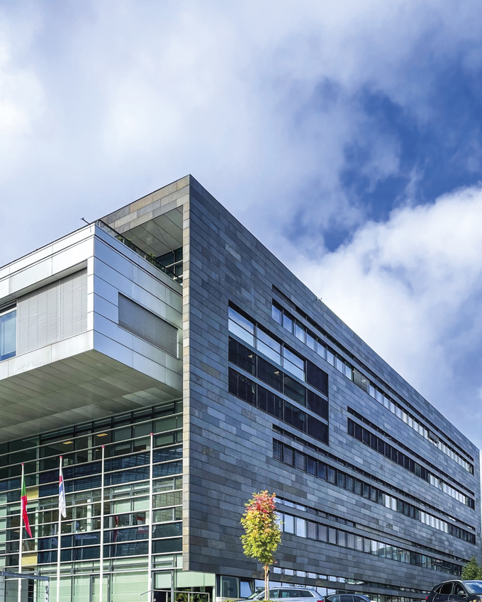 Moderne bygning med en elegant stenfacade og store glasvinduer under en delvis overskyet himmel. Træer og biler er i forgrunden.