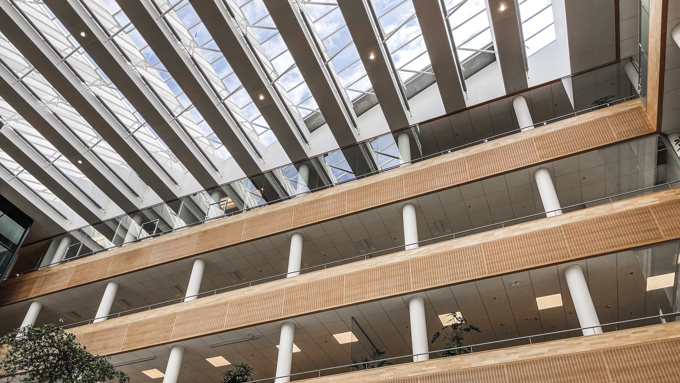 Indvendigt billede af en moderne bygning med træpaneler, store glasvinduer og grønne planter på flere niveauer.